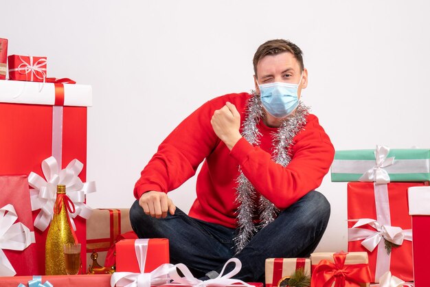 Vista frontale del giovane in maschera seduto intorno ai regali di Natale sul muro bianco