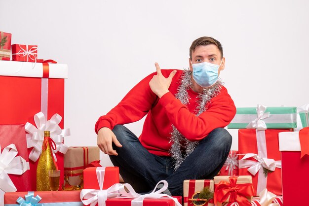 Vista frontale del giovane in maschera seduto intorno ai regali di Natale sul muro bianco
