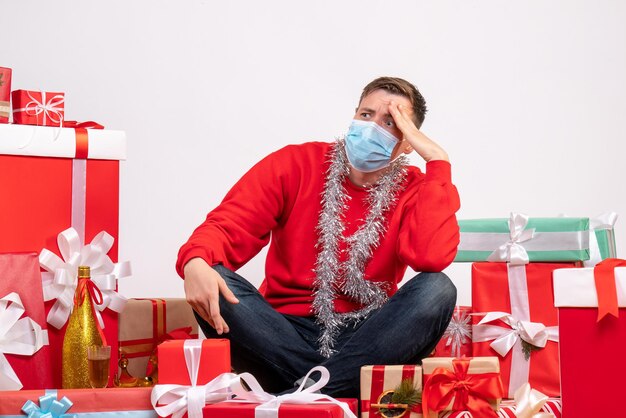 Vista frontale del giovane in maschera seduto intorno ai regali di Natale sul muro bianco