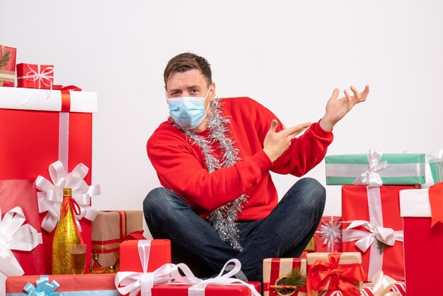 Vista frontale del giovane in maschera seduto intorno ai regali di Natale sul muro bianco