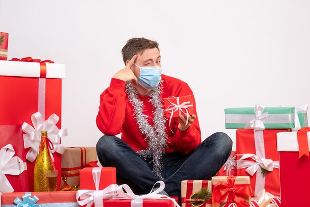 Vista frontale del giovane in maschera seduto intorno ai regali di Natale sul muro bianco