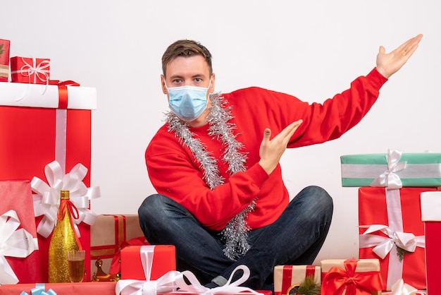 Vista frontale del giovane in maschera seduto intorno ai regali di Natale sul muro bianco