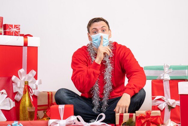 Vista frontale del giovane in maschera seduto intorno ai regali di Natale sul muro bianco