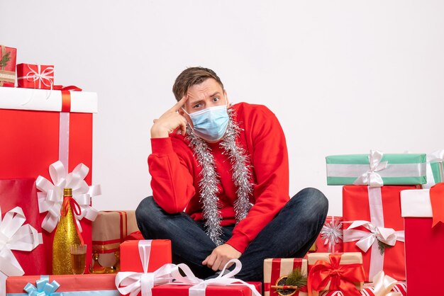 Vista frontale del giovane in maschera seduto intorno ai regali di Natale sul muro bianco