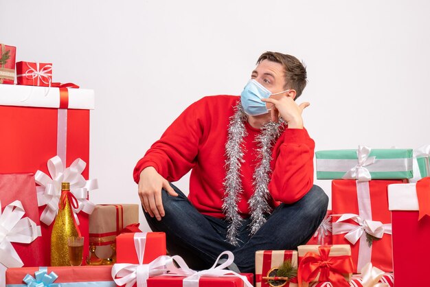 Vista frontale del giovane in maschera seduto intorno ai regali di Natale sul muro bianco