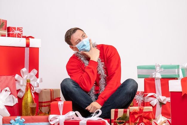 Vista frontale del giovane in maschera seduto intorno ai regali di Natale sul muro bianco