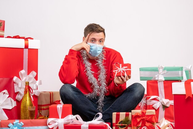 Vista frontale del giovane in maschera seduto intorno ai regali di Natale sul muro bianco
