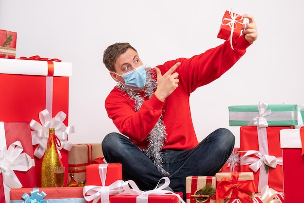 Vista frontale del giovane in maschera sterile seduto intorno ai regali di Natale sul muro bianco