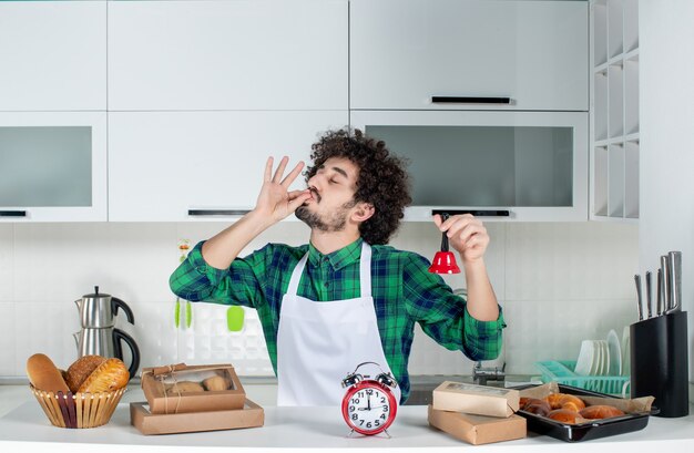 Vista frontale del giovane in piedi dietro il tavolo vari pasticcini su di esso e che mostra un campanello rosso che fa il gesto degli occhiali nella cucina bianca