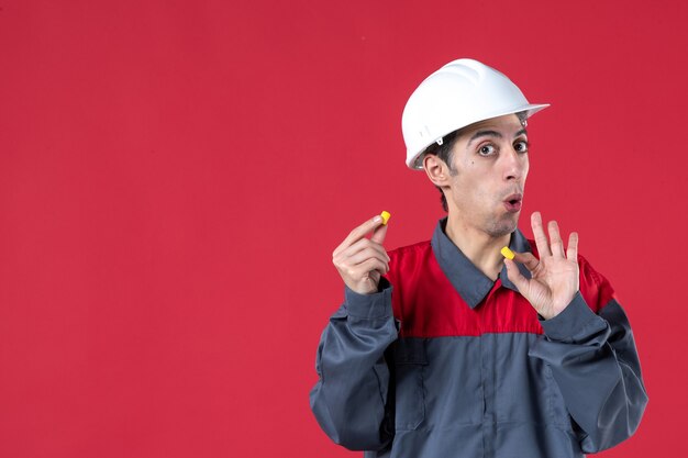 Vista frontale del giovane lavoratore interessato in uniforme con elmetto e tenendo i tappi per le orecchie sul muro rosso isolato