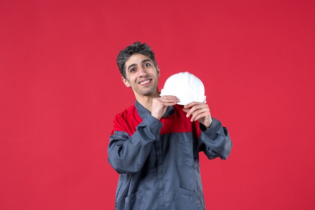 Vista frontale del giovane lavoratore sorridente in uniforme e che tiene il casco sul muro rosso isolato