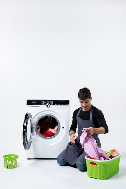 Vista frontale del giovane maschio che lava i vestiti con l'aiuto della lavatrice sul muro bianco