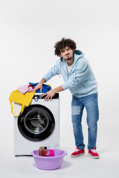 Vista frontale del giovane maschio con lavatrice e vestiti sporchi sul muro bianco