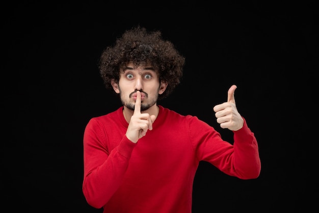 Vista frontale del giovane maschio in camicia rossa che chiede di tacere sul muro nero