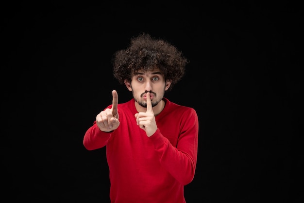 Vista frontale del giovane maschio in camicia rossa che chiede di tacere sul muro nero