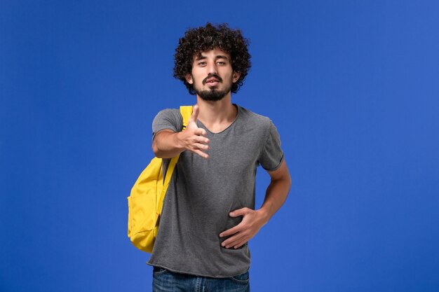 Vista frontale del giovane maschio in maglietta grigia che porta zaino giallo che prova a stringere la mano sulla parete blu