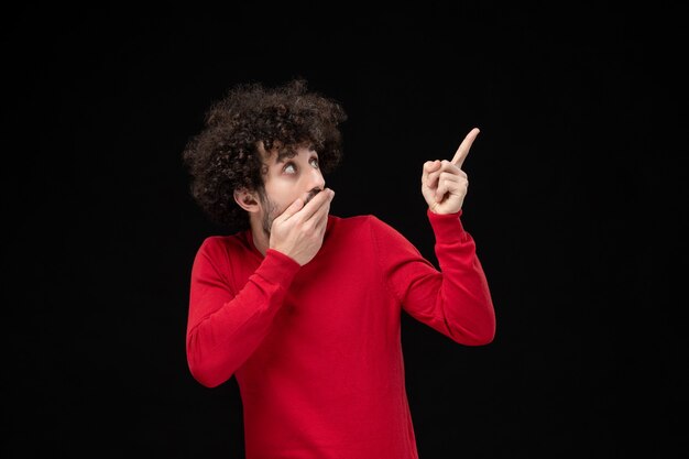 Vista frontale del giovane maschio in maglione rosso con faccia scioccata sul muro nero