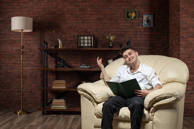 Vista frontale del giovane maschio sorridente che si siede sul sofà e che scrive le note all'interno dell'ufficio di lavoro della casa della mobilia del lavoro di affari della stanza