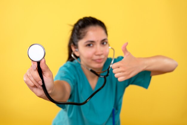 Vista frontale del giovane medico femminile con lo stetoscopio che fa il pollice sul segno sulla parete gialla