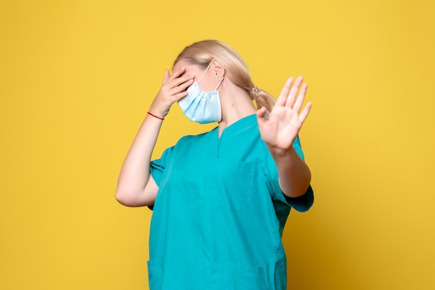 Vista frontale del giovane medico femminile in camicia medica e maschera sterile sulla parete gialla