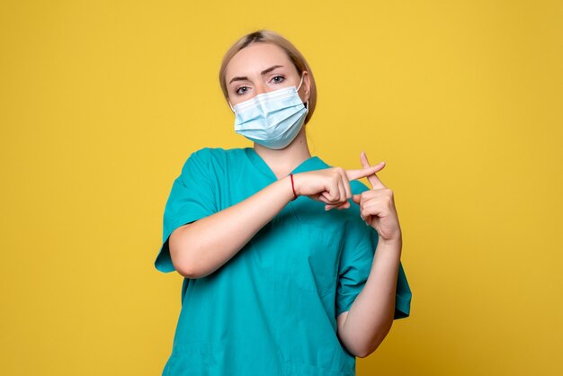 Vista frontale del giovane medico femminile in camicia medica e maschera sterile sulla parete gialla