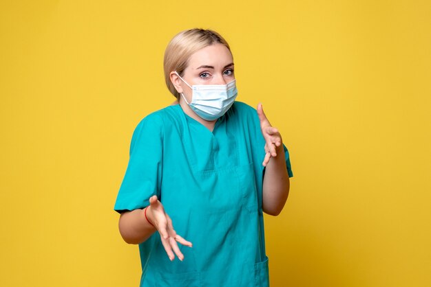 Vista frontale del giovane medico femminile in camicia medica e maschera sulla parete gialla