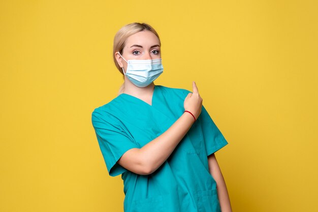 Vista frontale del giovane medico femminile in camicia medica e maschera sulla parete gialla