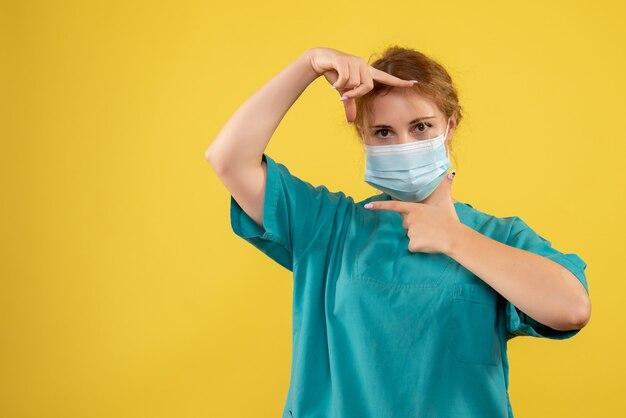 Vista frontale del giovane medico femminile in tuta medica e maschera sulla parete gialla