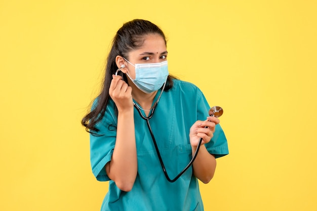 Vista frontale del giovane medico in uniforme utilizzando uno stetoscopio in piedi sulla parete gialla