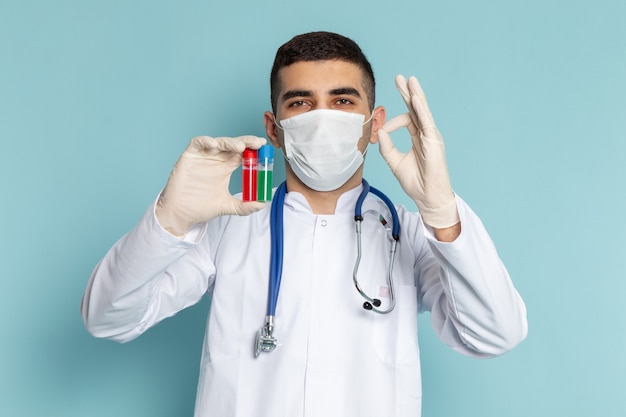 Vista frontale del giovane medico maschio in abito bianco con stetoscopio blu che tiene boccette con maschera