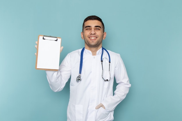 Vista frontale del giovane medico maschio in vestito bianco con il blocco note blu della tenuta dello stetoscopio