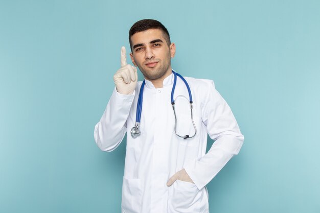 Vista frontale del giovane medico maschio in vestito bianco con la posa blu dello stetoscopio