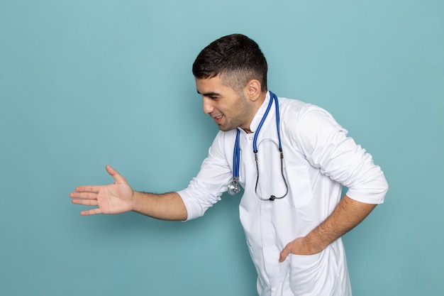 Vista frontale del giovane medico maschio in vestito bianco con lo stetoscopio blu che agita la mano