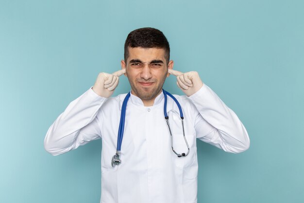 Vista frontale del giovane medico maschio in vestito bianco con lo stetoscopio blu che colpisce le sue orecchie