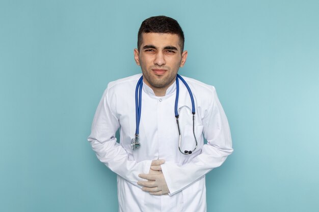 Vista frontale del giovane medico maschio in vestito bianco con lo stetoscopio blu che ha mal di stomaco