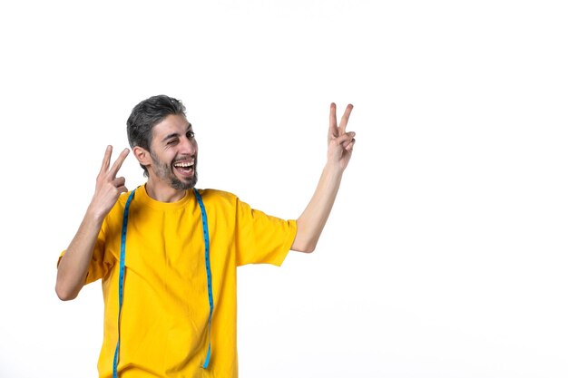 Vista frontale del giovane ragazzo sorridente in camicia gialla e tenendo il misuratore che fa il gesto di vittoria sulla superficie bianca