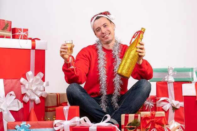 Vista frontale del giovane seduto intorno ai regali che versano champagne sul muro bianco