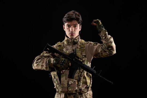 Vista frontale del giovane soldato in uniforme con il fucile sul muro nero