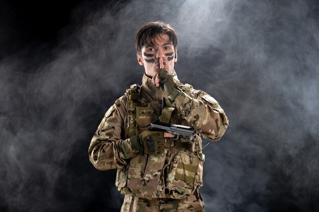 Vista frontale del giovane soldato in uniforme con la pistola sul muro nero