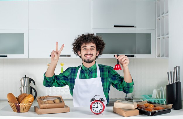 Vista frontale del giovane sorridente in piedi dietro il tavolo vari pasticcini su di esso e che mostra campanello rosso che fa gesto di vittoria nella cucina bianca