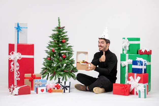 Vista frontale del giovane uomo seduto intorno ai regali di festa che tiene il pacchetto con i giocattoli sul muro bianco