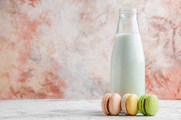 Vista frontale del latte fresco in una bottiglia aperta accanto a deliziosi macarons colorati sul lato sinistro sulla superficie dei colori pastello