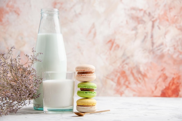 Vista frontale del latte fresco in una tazza di vetro e bottiglia accanto a deliziosi macarons colorati fiori cucchiaio d'oro sul lato destro su sfondo colori pastello