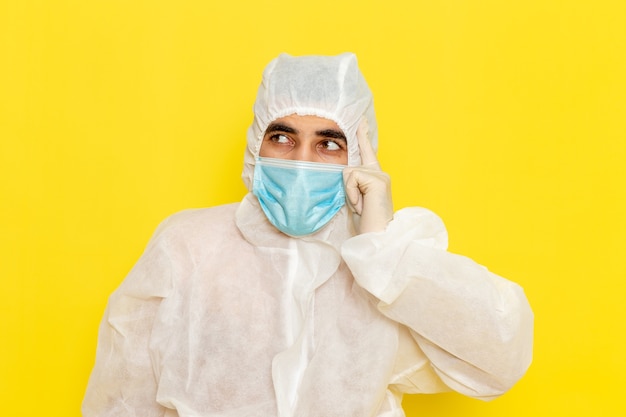 Vista frontale del lavoratore scientifico maschio in abito bianco protettivo speciale con maschera sterile pensando sulla parete gialla