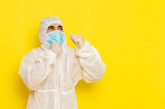 Vista frontale del lavoratore scientifico maschio in tuta protettiva speciale e con la maschera in posa sulla parete giallo chiaro