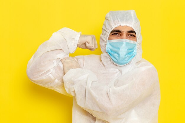 Vista frontale del lavoratore scientifico maschio in tuta protettiva speciale e con maschera che flette sul muro giallo chiaro