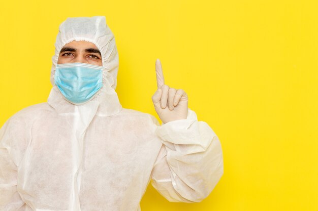 Vista frontale del lavoratore scientifico maschio in vestito bianco protettivo speciale con maschera sterile alzando il dito sulla foto di pericolo colore chimica chimica operaio di scienza scrivania gialla