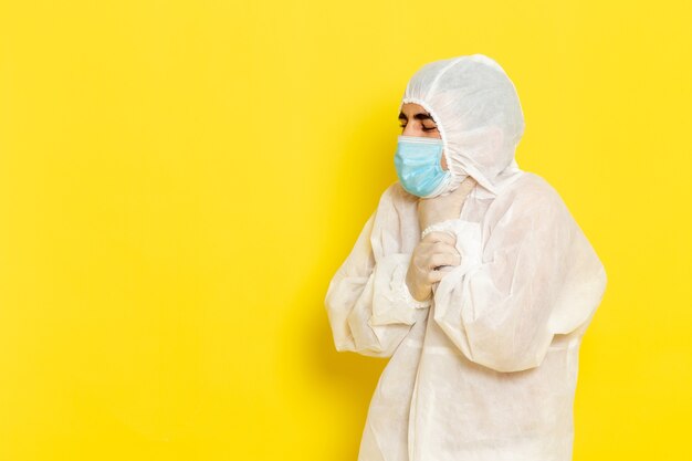 Vista frontale del lavoratore scientifico maschio in vestito bianco protettivo speciale e con la maschera che ha problemi alla gola sulla parete gialla