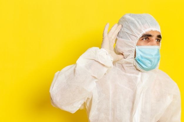 Vista frontale del lavoratore scientifico maschio in vestito bianco protettivo speciale e con la maschera che prova a sentire sulla parete gialla chiara