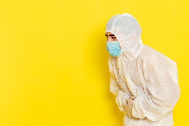 Vista frontale del lavoratore scientifico maschio in vestito bianco protettivo speciale e con la maschera che tiene il suo stomaco sulla foto di pericolo di colore di chimica del lavoratore di scienza della scrivania gialla